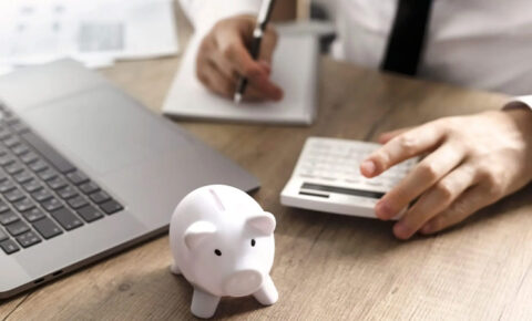 Berater berechnet Finanzen mit Taschenrechner und Laptop, weißes Sparschwein im Vordergrund auf einem Holztisch