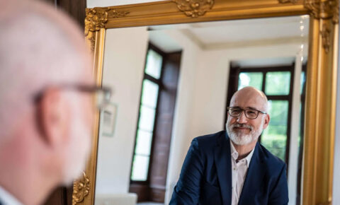 Mann mit Brille und Blazer betrachtet sich in einem vergoldeten Spiegel